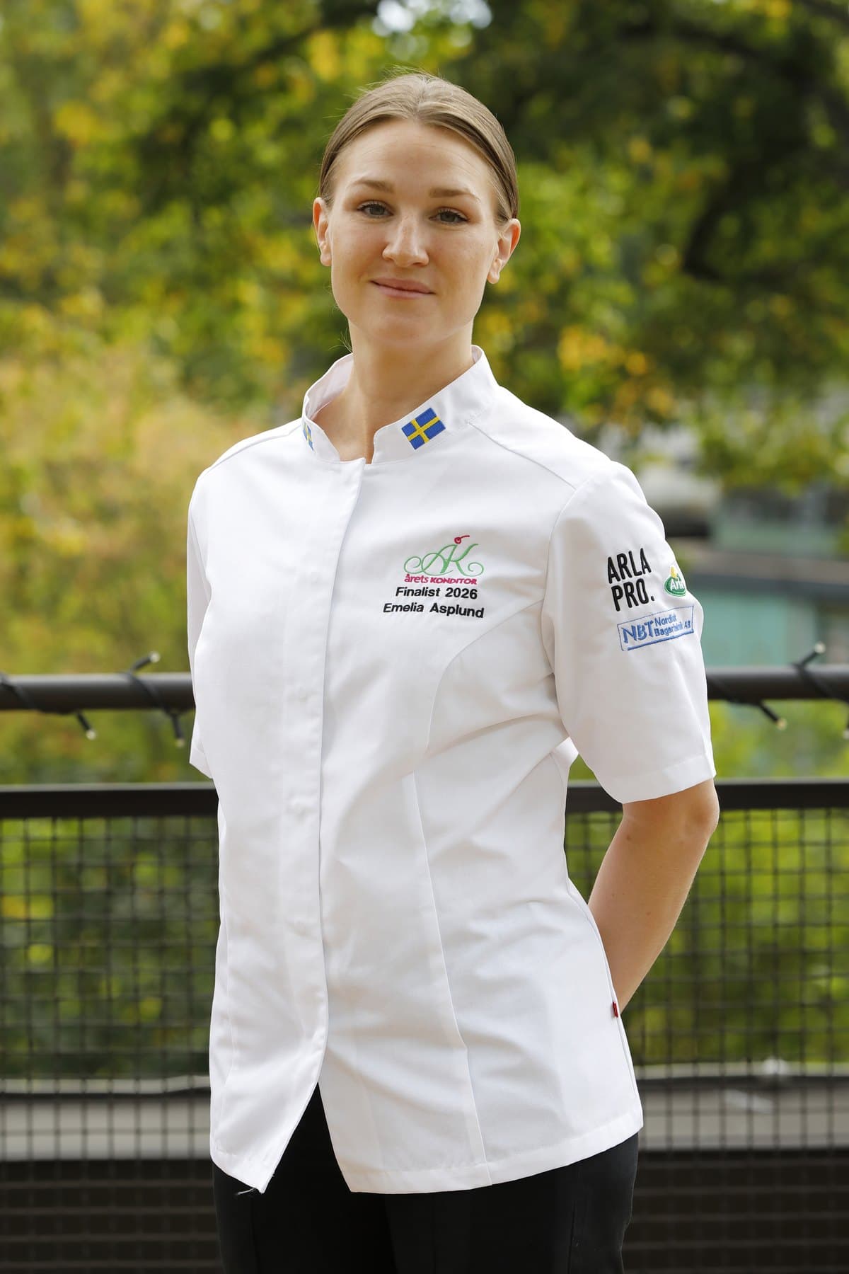 Emelia Asplund i vit kockrock med texten Årets Konditor, Finalist 2026, utomhus i höstljus